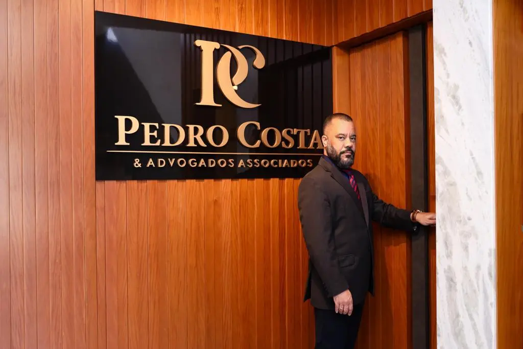 Dr. Pedro Costa diante da placa do escritório, no painel de madeira da recepção